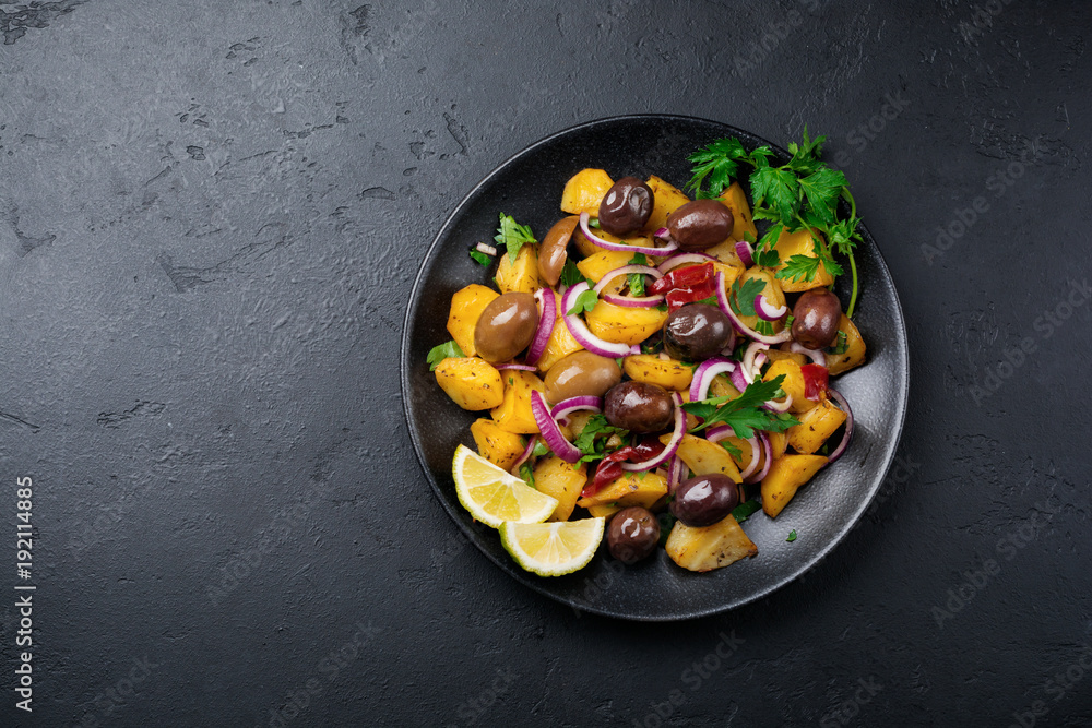 Warm potato salad with olives, pepper, parsley and red onion on black ceramic plate on dark concrete background. Selective focus. Top view. Copy space.