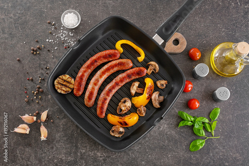Grill pan with delicious sausages and vegetables on grey background