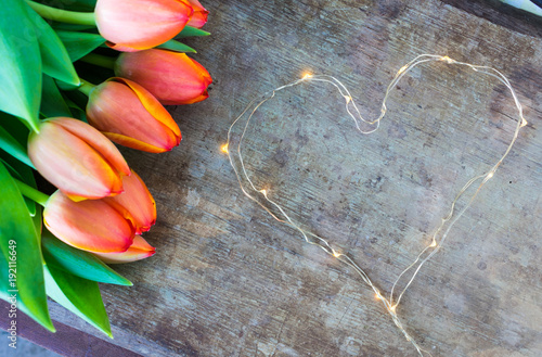 View of colorful orange tulips and led lights chain shaped in heart form on old rustic vintage wooden background. Love, valentines or mother's day background #192116649