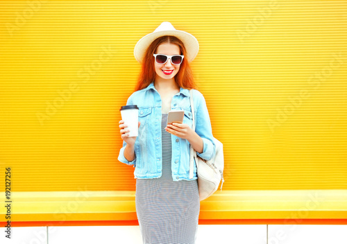 Fashion smiling woman using a smartphone holds coffee cup on a orange background