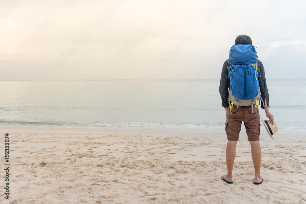 Young Asian backpacker man standing on the beach and holding hat, summer holiday vacation and travel tropical island concepts