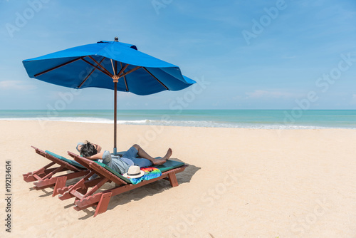 Young Asian man traveler lying on beach bench with blue umbrella overhead. surrounded by beautiful tropical beach island and blue sky, vacation time and summer holiday for digital nomad lifestyle