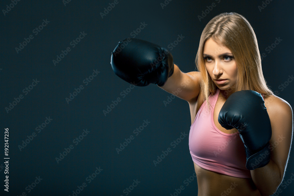 Woman in boxing gloves and sportswear strikes at workout on dark background. Copy space