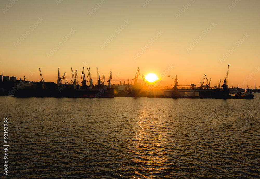 Industrial photo of busy port in the city. Sunset over the sea and cargo ships. Yellow sun and deep water. Romantic photo harbor.