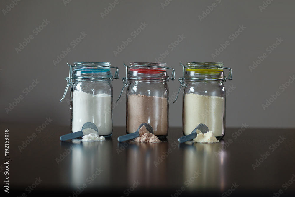 Three glass jars with protein powder, strawberries, banana and chocolate and three scoops for milkshakes. Sports and dietary nutrition for athletes and weight loss or muscle mass bodybuilders. Three glass jars with protein powder, strawberries, banana and chocolate and three scoops for milkshakes. Sports and dietary nutrition for athletes and weight loss or muscle mass bodybuilders.