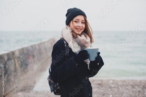 Young beautiful teenager girl drinks coffee and smiles on the beach in winter.
