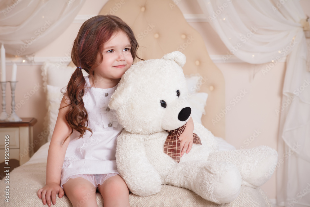 A little girl in her room sits on a bed and holds a bear. A child in a white dress with a toy. Beige interior.