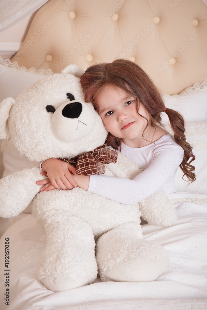 A little girl lies in a bed in a baby's beige room. He hugs a toy of a polar bear. Good morning.