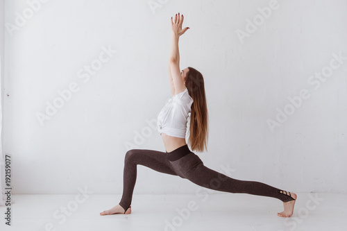 Side view portrait of beautiful young woman wearing white tank top working out against white wall, doing yoga or pilates exercise. Standing in Warrior one pose, Virabhadrasana. Full length