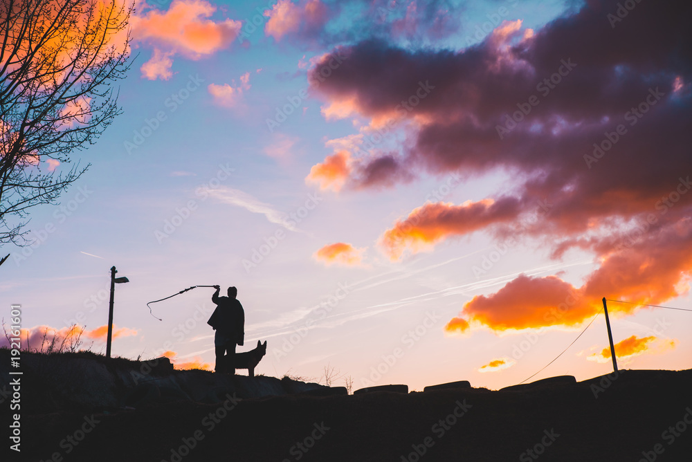 Man with whip and dog, silhouette photo, sunset sky in background