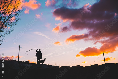 Man with whip and dog, silhouette photo, sunset sky in background