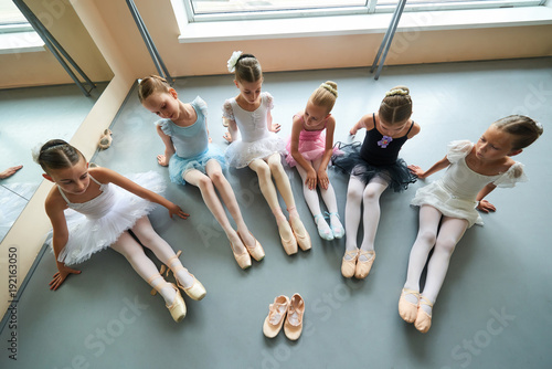 Six ballerinas sitting on floor, top view. Pair of slippers for little ballet girl. Group of young ballet dancers in different dresses.