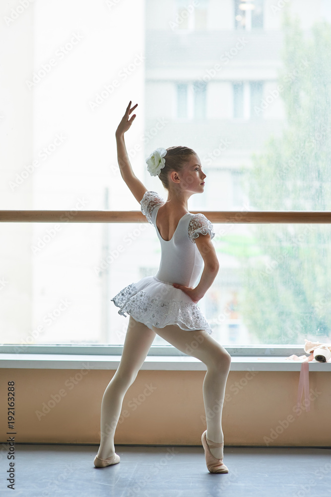 Young ballerina standing in ballet position. Beautiful young ballet girl in white dress posing in ballet dance class. Talented and skillful youth.