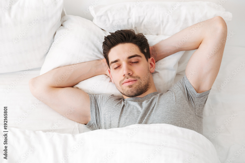 View from above of brunette muscular man in casual t-shirt, sleeping alone at home in bed with white clean linen putting hands under head View from above of brunette muscular man in casual t-shirt, sleeping alone at home in bed with white clean linen putting hands under head