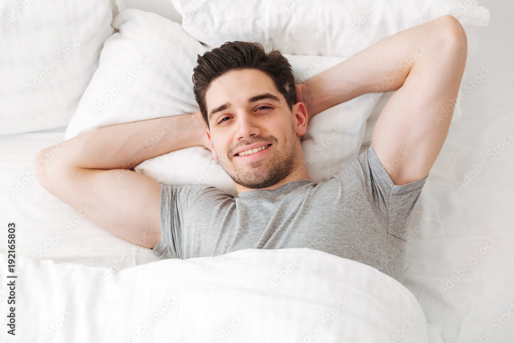 View from above of brunette sexual man in casual t-shirt smiling on camera while lying alone at home in white bed, with putting hands under head View from above of brunette sexual man in casual t-shirt smiling on camera while lying alone at home in white bed, with putting hands under head