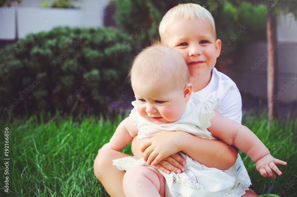 happy little brother playing hugs his sister baby sitting on grass in a green garden, concept of love and parenting