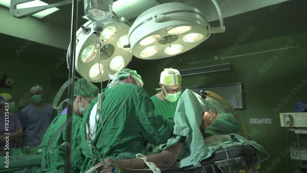 Elderly man lying on table under anesthesia with oxygen mask on head and intravenous drip injected in hand during surgeons team using surgical tools to operating patient at operation room, dolly shot Elderly man lying on table under anesthesia with oxygen mask on head and intravenous drip injected in hand during surgeons team using surgical tools to operating patient at operation room, dolly shot