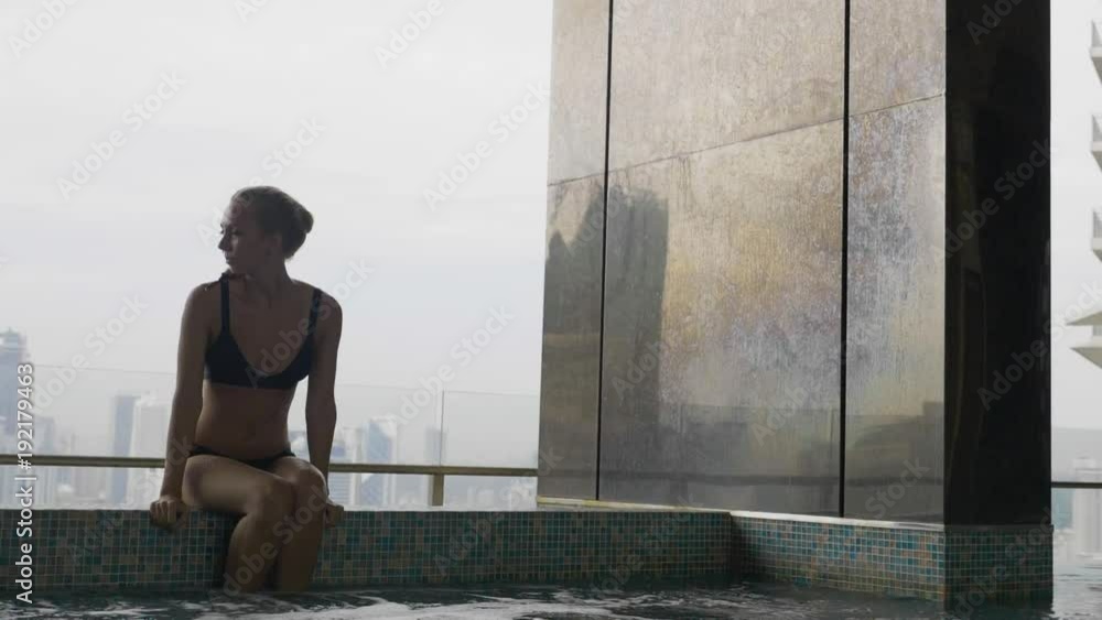 Pretty slim rich blonde woman in black bikini relaxing at the edge of roof top swimming pool or jakuzzi at resort or in hotel with view over city skyline. Success, vacation leisure activity concept. Pretty slim rich blonde woman in black bikini relaxing at the edge of roof top swimming pool or jakuzzi at resort or in hotel with view over city skyline. Success, vacation leisure activity concept.