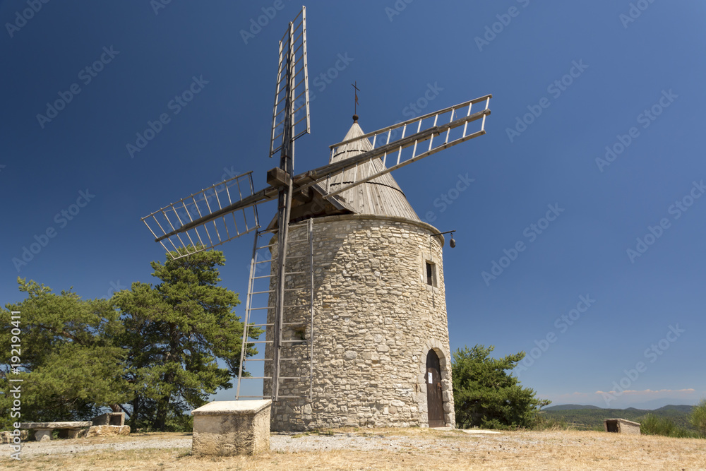 Old stone windmill and small group of green trees from one side with clear blue sky in Provence, France  