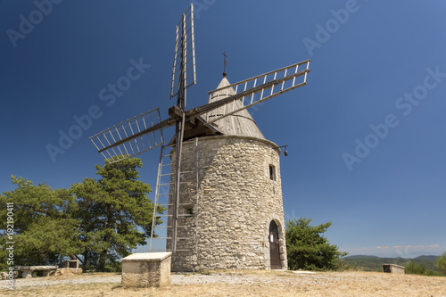 Old stone windmill and small group of green trees from one side with clear blue sky in Provence, France  
