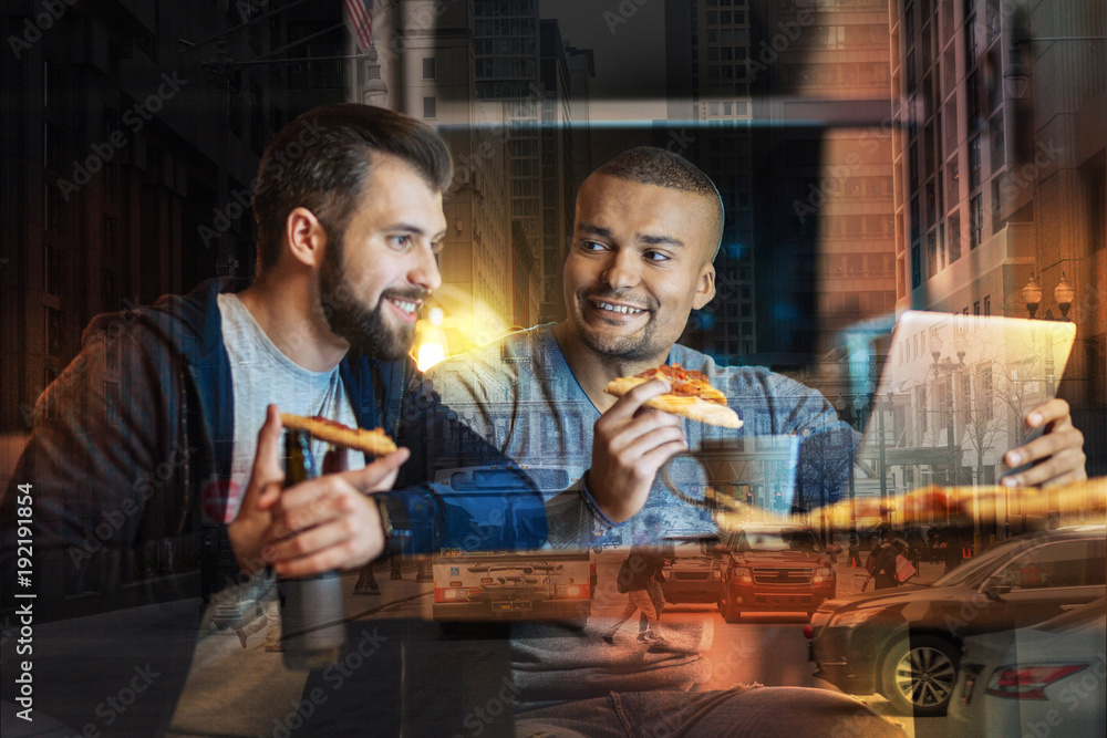 Tasty food. Bearded young man eating pizza and looking at the screen of a laptop while his cheerful friend sitting near and looking attentively at him