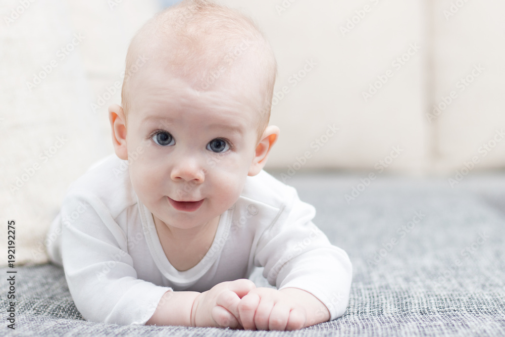 Cute, smiling, crawling baby boy on grey couch, wearing white dress.
