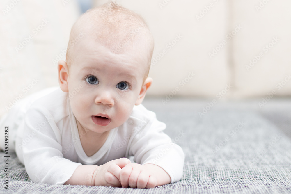 Cute, smiling, crawling baby boy on grey couch, wearing white dress.