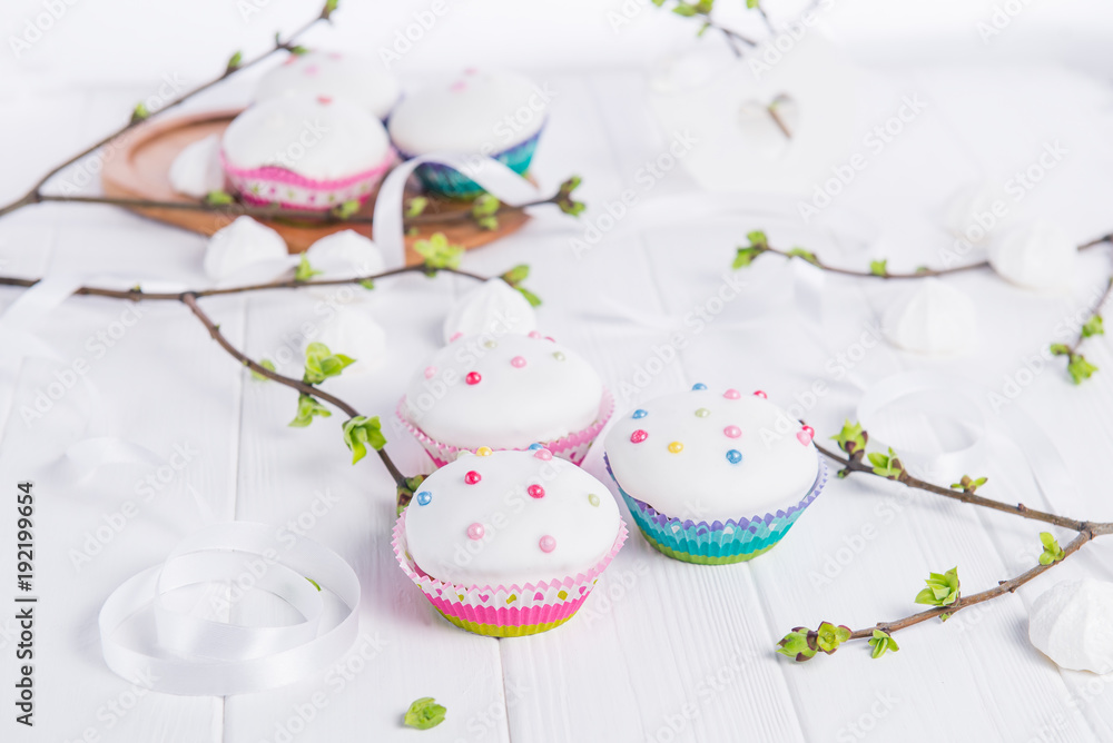 Decorated Easter cupcakes on the white wooden table with branches with young shoots of greenery, satin ribbon and light merengue sweets. Holiday concept. Selective focus, copy space.