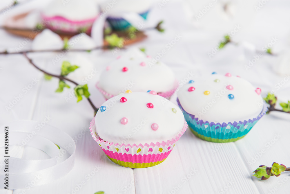 Decorated Easter cupcakes on the white wooden table with branches with young shoots of greenery, satin ribbon and light merengue sweets. Holiday concept. Selective focus, copy space. Decorated Easter cupcakes on the white wooden table with branches with young shoots of greenery, satin ribbon and light merengue sweets. Holiday concept. Selective focus, copy space.