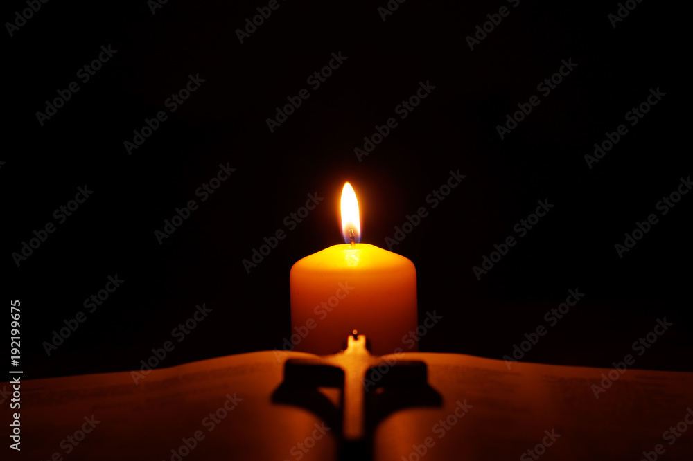 Burning candle, open Bible and crucifix in the dark.