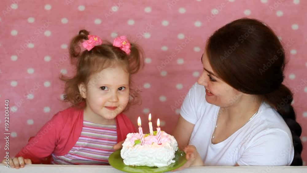 Happy mom and child make a wish and blows out candles on birthday cake at party. Mother Congratulates, embraces and Kisses Her Daughter on a pink background. 3 years Happy mom and child make a wish and blows out candles on birthday cake at party. Mother Congratulates, embraces and Kisses Her Daughter on a pink background. 3 years