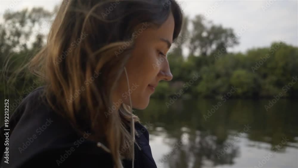 Side view of a young woman wearing a black hoodie listening to the music and walking along a river bank. Tracking real time close up shot Side view of a young woman wearing a black hoodie listening to the music and walking along a river bank. Tracking real time close up shot