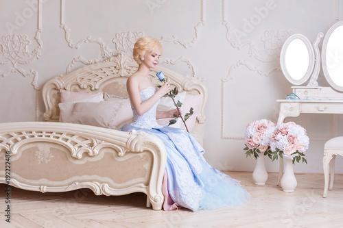 Fényképezés A beautiful, young blonde woman sits on a bed in a bedroom, dressed in a ball gown, and holds a blue rose in her hands