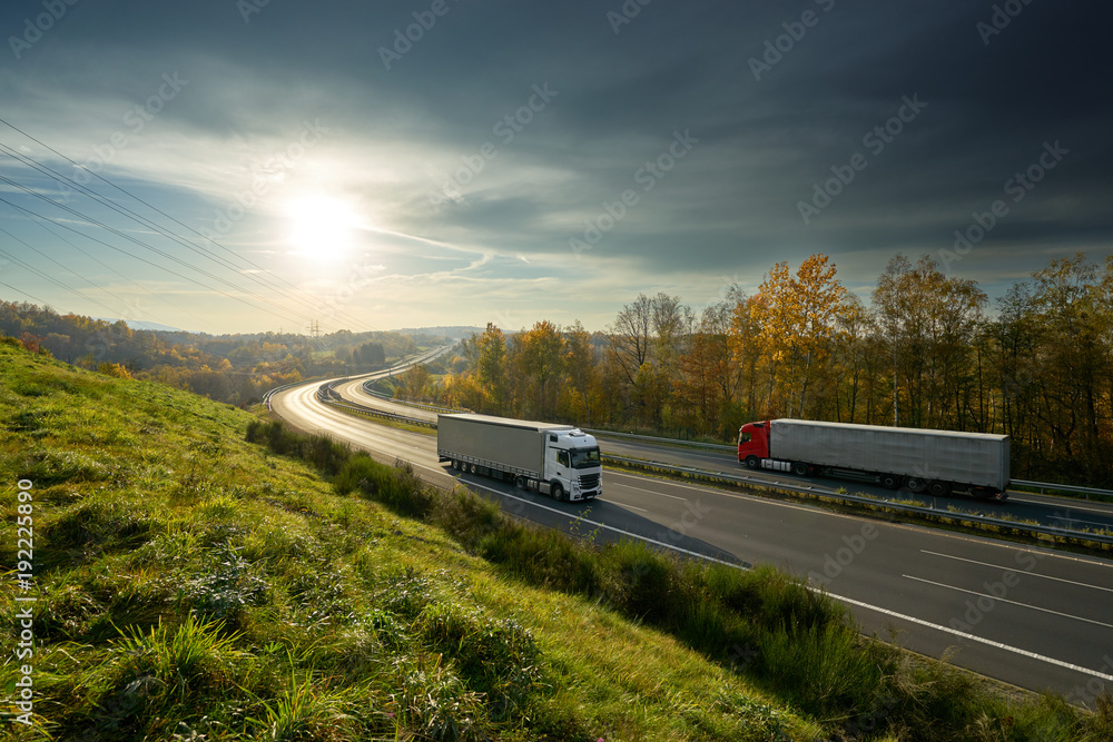 Trucks driving on the highway turning towards the horizon in an autumn landscape with sun shining through the clouds in the sky