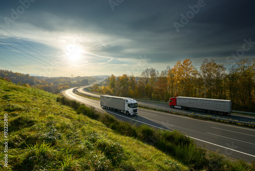 Obraz na płótnie Trucks driving on the highway turning towards the horizon in an autumn landscape with sun shining through the clouds in the sky w salonie