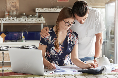 Family budget, finances and people concept. Handsome male embraces his beautiful young wife, work with documents and calculate bills in kitchen, pay taxes on laptop computer. Domestic atmosphere photo