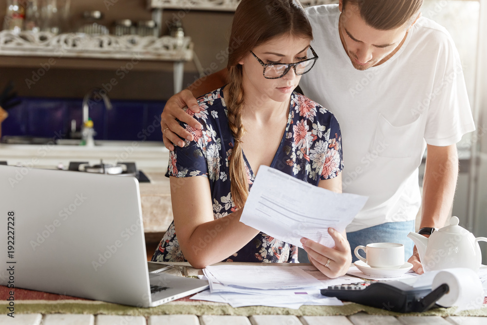 Cropped view of attractive young female and man study bank papers attentively, going to take loan for buying new lodging, have many debts, pose against kitchen interior with laptop computer. Cropped view of attractive young female and man study bank papers attentively, going to take loan for buying new lodging, have many debts, pose against kitchen interior with laptop computer.