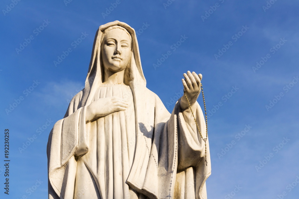 Virgin Mary statue holding rosary in hand with blue clear sky background stands in front of the Roman Catholic Church Pfarrkirche St. Alban in Matrei in Osttirol
