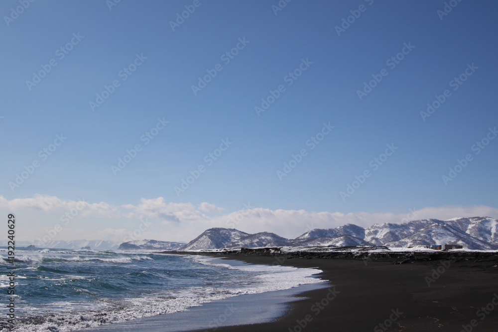 Beautiful winter seascape of Pacific ocean with big waves, black volcano sand and snowy mountains background. View of Avacha bay on Kamchatka Peninsula nearby the city Petroplavlovsk-Kamchatsky.