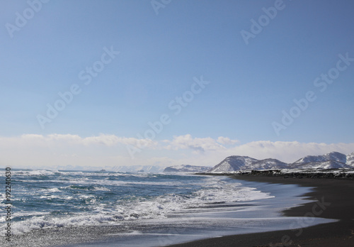 Beautiful winter seascape of Pacific ocean with big waves, black volcano sand and snowy mountains background. View of Avacha bay on Kamchatka Peninsula nearby the city Petroplavlovsk-Kamchatsky.