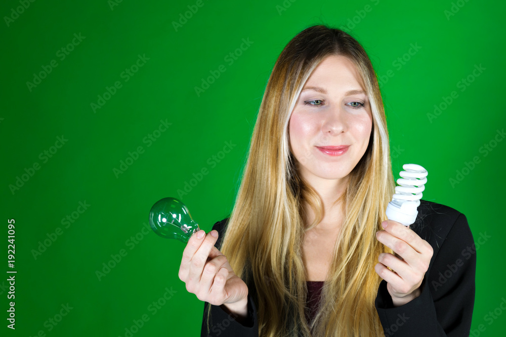 Serious pretty business woman holding a light bulb on a green background. Close-up face model with two kinds of light bulbs. Concept of saving money for electricity Serious pretty business woman holding a light bulb on a green background. Close-up face model with two kinds of light bulbs. Concept of saving money for electricity