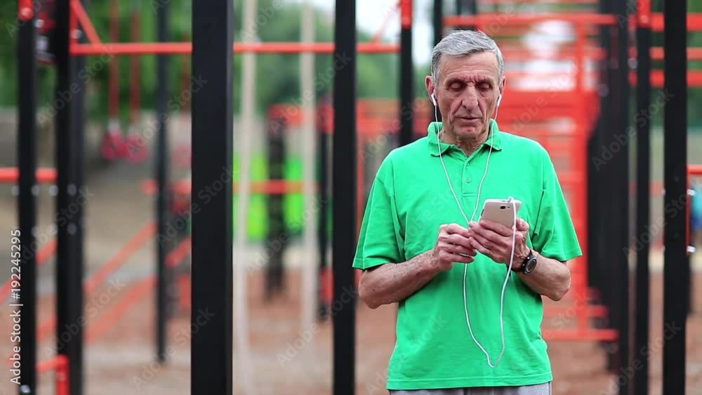 Senior man with cell phone stands near sports ground. Active elderly athlete hold in hands mobile phone. Aged sportsman listens to music on smartphone. Grey-haired man in the open air gym Senior man with cell phone stands near sports ground. Active elderly athlete hold in hands mobile phone. Aged sportsman listens to music on smartphone. Grey-haired man in the open air gym