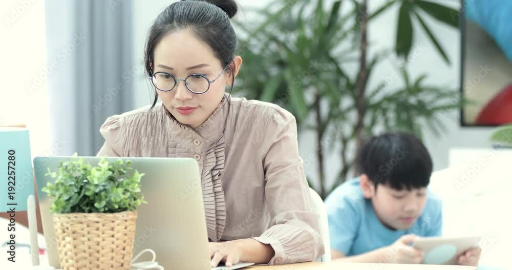 Shot of an attractive mature businesswoman working on laptop in her workstation at home with boy playing tablet computer on background. Shot of an attractive mature businesswoman working on laptop in her workstation at home with boy playing tablet computer on background.