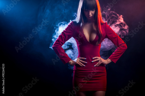 Young dark-haired woman in a red dress posing against a background of red and  blue smoke from a vape on a black isolated background