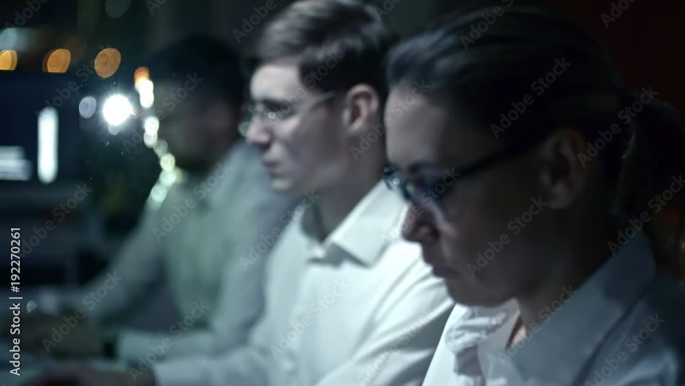 Rack focused shot of IT professionals working on computers in dark office at night with projector lightning at camera in background Rack focused shot of IT professionals working on computers in dark office at night with projector lightning at camera in background