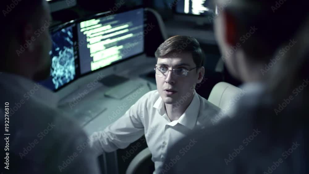 Tracking shot of young IT professional programmer in eyeglasses working on computer in cyber security center filled with display screens and talking with colleagues Tracking shot of young IT professional programmer in eyeglasses working on computer in cyber security center filled with display screens and talking with colleagues