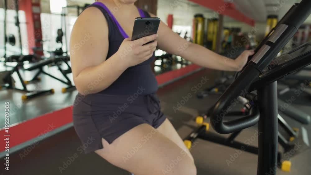 Overweight woman riding slowly stationary bike and chatting on mobile phone Overweight woman riding slowly stationary bike and chatting on mobile phone