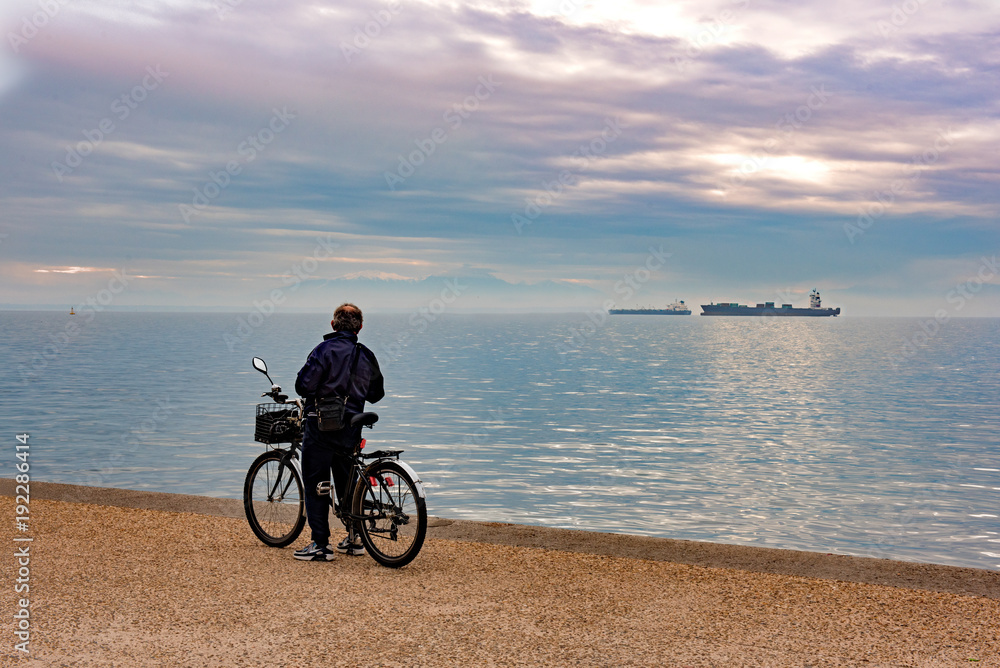 Fototapeta premium Senior man with his bicycle standing and enjoy the view along the seafront in the sunrise. Thessaloniki, Greece