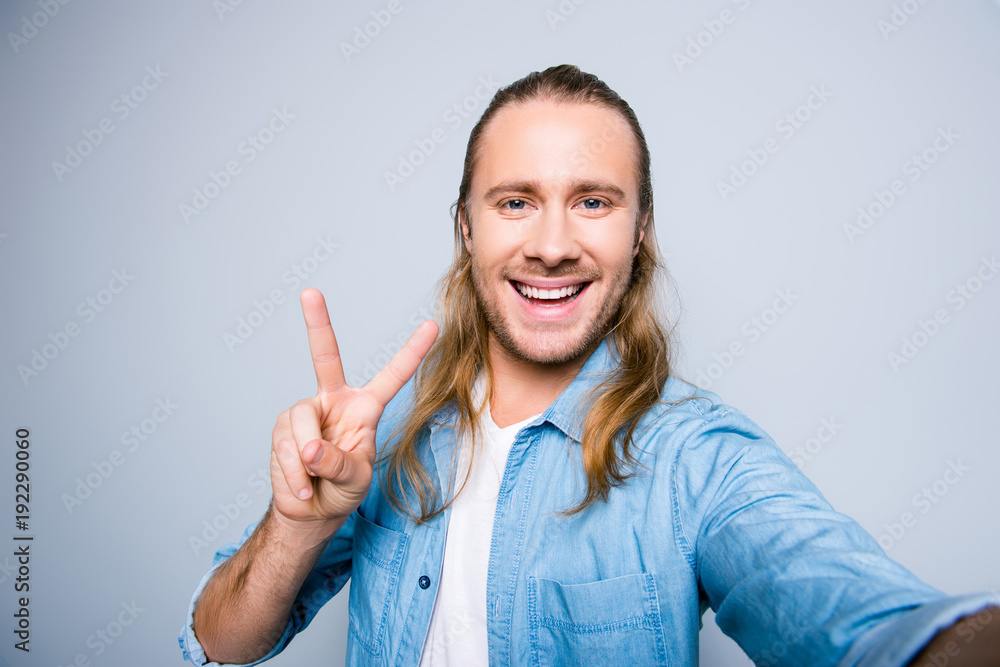Close up picture of attractive, bearded blogger with long hair shooting selfie, gesturing peace symbol with two fingers, having life video chat, standing over grey background Close up picture of attractive, bearded blogger with long hair shooting selfie, gesturing peace symbol with two fingers, having life video chat, standing over grey background