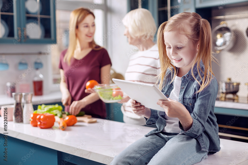 Great pastime. Pretty little girl sitting on the kitchen counter and playing on the tablet while her mother and grandmother cooking lunch in the background Great pastime. Pretty little girl sitting on the kitchen counter and playing on the tablet while her mother and grandmother cooking lunch in the background